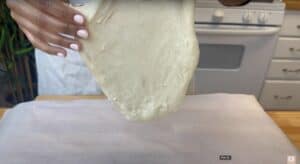 Person stretching pizza dough over parchment paper in a kitchen setting.